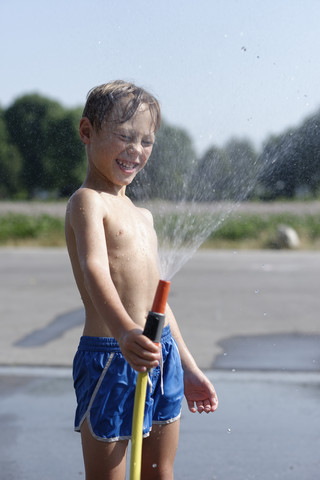 Lachender Junge, der im Sommer mit einem Gartenschlauch Wasser verspritzt, lizenzfreies Stockfoto