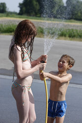 Lachender Junge bespritzt seine Schwester mit einem Gartenschlauch im Sommer mit Wasser - LBF001325