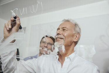 Two men writing onto glass wall in office - MFF002537