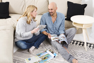 Ein Paar sitzt auf dem Boden seines Wohnzimmers und sieht sich Reisekataloge an - MAEF011167