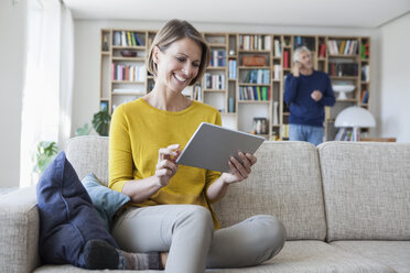 Lächelnde Frau sitzt auf der Couch und benutzt ein digitales Tablet, während ihr Mann im Hintergrund telefoniert - RBF003763