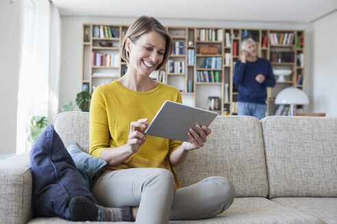 Smiling woman sitting on the couch using digital tablet while her husband telephoning in the background - RBF003763