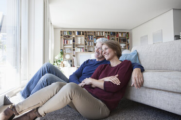 Ein glückliches Paar sitzt Seite an Seite auf dem Boden ihres Wohnzimmers - RBF003768