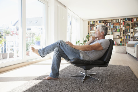 Nachdenklicher Mann entspannt sich auf einem Ledersessel zu Hause und schaut durch das Fenster, lizenzfreies Stockfoto
