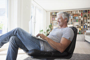 Lächelnder Mann entspannt sich auf einem Ledersessel zu Hause und benutzt einen Laptop - RBF003800