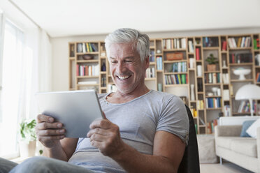Porträt eines lachenden Mannes, der im Wohnzimmer sitzt und auf ein digitales Tablet schaut - RBF003801