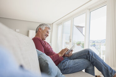 Lächelnder Mann, der zu Hause auf der Couch sitzt und ein digitales Tablet benutzt - RBF003807