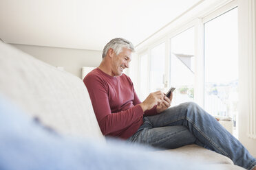 Lächelnder Mann, der zu Hause auf der Couch sitzt und sein Smartphone benutzt - RBF003809