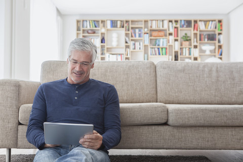 Lächelnder Mann, der zu Hause vor der Couch sitzt und ein digitales Tablet benutzt, lizenzfreies Stockfoto