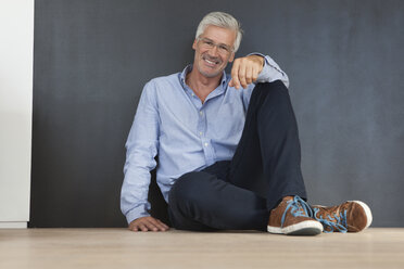 Portrait of smiling mature man sitting on the floor in front of a grey wall - RBF003828