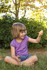 Smiling little girl sitting on a meadow watching something in her hand - LVF004334