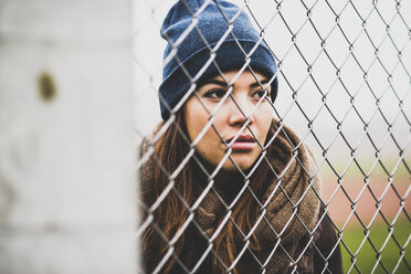 Portrait of young woman wearing wool cap looking through wire mesh - UUF006247