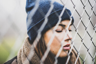 Portrait of young woman with closed eyes wearing wool cap behind wire mesh - UUF006250