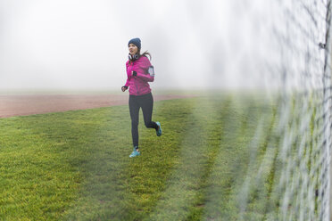 Junge Frau joggt auf einer Wiese hinter Maschendrahtzaun - UUF006253