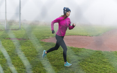 Junge Frau mit Kopfhörern joggt auf einer Wiese hinter Maschendraht - UUF006255