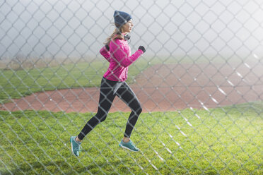 Junge Frau joggt auf einer Wiese hinter Maschendrahtzaun - UUF006260