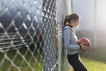 Young woman with basketball leaning against mesh wire fence on a sports field - UUF006265