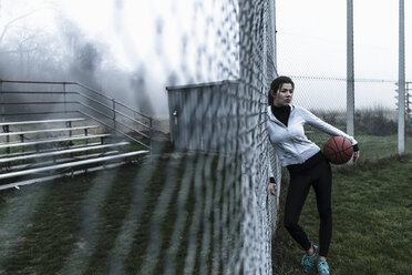 Junge Frau mit Basketball, der an einem Maschendrahtzaun auf einem Sportplatz lehnt - UUF006266