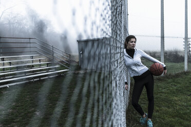 Young woman with basketball leaning against mesh wire fence on a sports field - UUF006266
