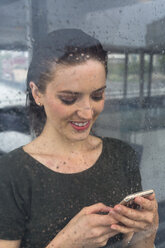 Lächelnde junge Frau hinter einer Fensterscheibe, die an einem regnerischen Tag auf ihr Smartphone schaut - GIOF000618