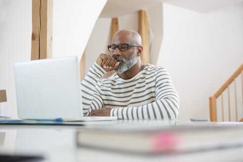 Portrait of man sitting at desk of home office looking at his laptop - RHF001152