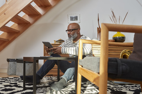 Mann sitzt auf dem Boden seines Wohnzimmers und benutzt ein digitales Tablet, lizenzfreies Stockfoto