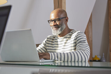 Porträt eines lächelnden Mannes, der an seinem Schreibtisch sitzt und mit einem Laptop arbeitet - RHF001190