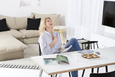 Laughing woman sitting at the table in her living room with magazine - MAEF011226