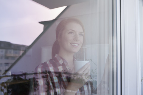 Porträt einer lächelnden jungen Frau mit einer Tasse Kaffee, die durch ein Fenster schaut, lizenzfreies Stockfoto