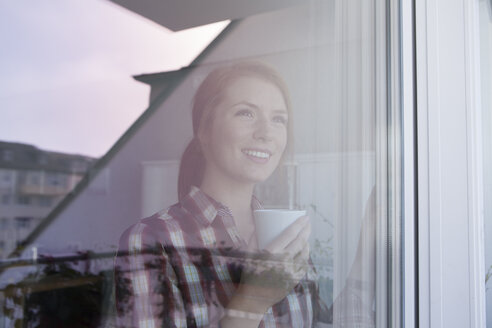 Porträt einer lächelnden jungen Frau mit einer Tasse Kaffee, die durch ein Fenster schaut - RBF003834