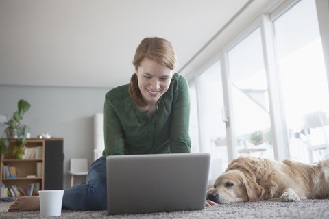 Lächelnde junge Frau sitzt neben ihrem Hund auf dem Teppich und benutzt einen Laptop, lizenzfreies Stockfoto