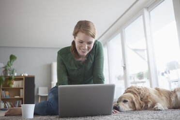 Lächelnde junge Frau sitzt neben ihrem Hund auf dem Teppich und benutzt einen Laptop - RBF003862