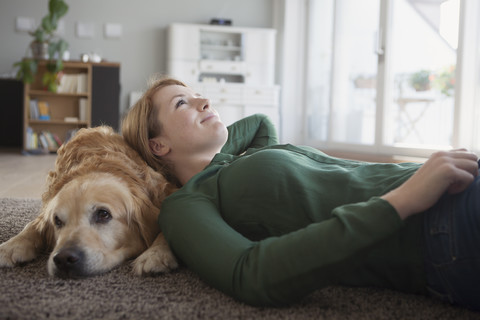 Lächelnde junge Frau, die mit ihrem Hund zu Hause auf dem Teppich liegt, lizenzfreies Stockfoto