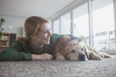 Lächelnde junge Frau liegt neben ihrem Hund auf dem Teppich - RBF003869