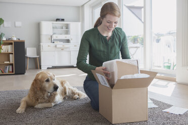 Lächelnde junge Frau, die neben ihrem Hund auf dem Boden kniet und ein Paket auspackt - RBF003870