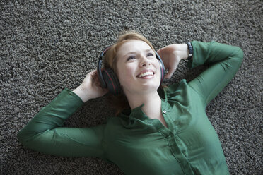 Lächelnde junge Frau, die auf einem Teppich liegt und mit Kopfhörern Musik hört - RBF003872