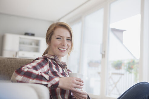 Porträt einer lächelnden jungen Frau, die sich mit einer Tasse Kaffee zu Hause entspannt - RBF003887