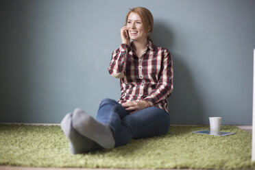 Lächelnde junge Frau, die auf dem Boden sitzt und mit ihrem Smartphone telefoniert - RBF003894