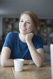 Porträt einer lächelnden jungen Frau, die am Tisch sitzt und eine Tasse Kaffee trinkt - RBF003896