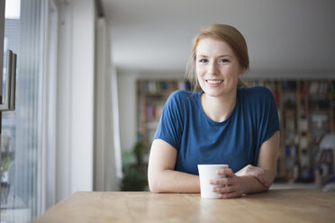 Porträt einer lächelnden jungen Frau, die am Tisch sitzt und eine Tasse Kaffee trinkt - RBF003899