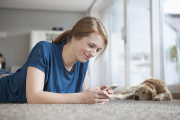 Lächelnde junge Frau, die zu Hause auf dem Boden liegt und auf ihr Smartphone schaut - RBF003905