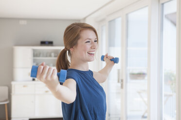 Portrait of smiling young woman exercising with dumbbells at home - RBF003908