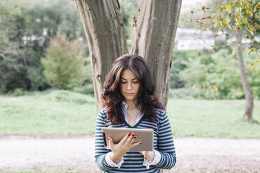 Junge Frau lehnt an einem Baumstamm und schaut auf ein digitales Tablet - ALBF000002