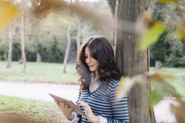 Smiling young woman leaning against tree trunk looking at digital tablet - ALBF000003
