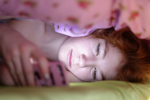 Portrait of girl lying under blanket in bed looking at her smartphone - LBF001335