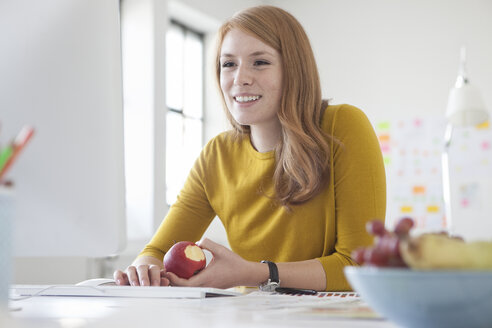 Junge Frau im Büro, die am Schreibtisch arbeitet und einen Apfel isst - RBF003944