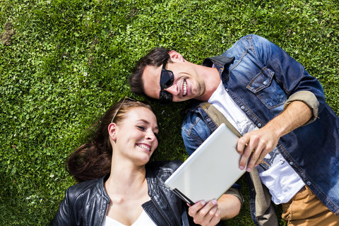 Glückliches junges Paar liegt auf einer Wiese und schaut auf ein digitales Tablet, lizenzfreies Stockfoto