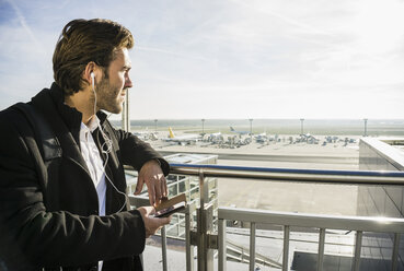 Germany, Frankfurt, Young businessman at the airport using smartphone with head phones - UUF006329