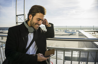 Germany, Frankfurt, Young businessman at the airport using smartphone with head phones - UUF006333