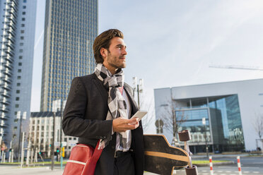 Germany, Frankfurt, Young businessman walking the city with skateboard, using mobile phone - UUF006334